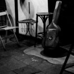 Fotografía de sala de conciertos. Hay una funda de guitarra sobre un taburete en lo que es el pequeño escenario y sillas de madera alrededor.