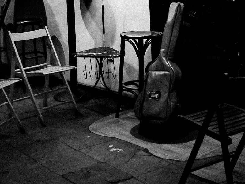 Fotografía de sala de conciertos. Hay una funda de guitarra sobre un taburete en lo que es el pequeño escenario y sillas de madera alrededor.