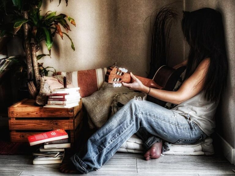 Mujer con una guitarra apoyada en la pared. Está en el rincón de una habitación junto a una mesa llena de libros.