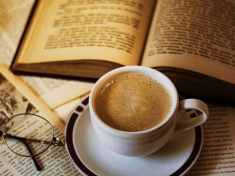 Taza de café sobre papeles. Hay un libro abierto y unas gafas al lado de la taza.