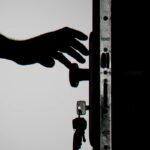 Mano de una persona en el picaporte de una puerta. Se ven las llaves puestas en la cerradura. En blanco y negro.