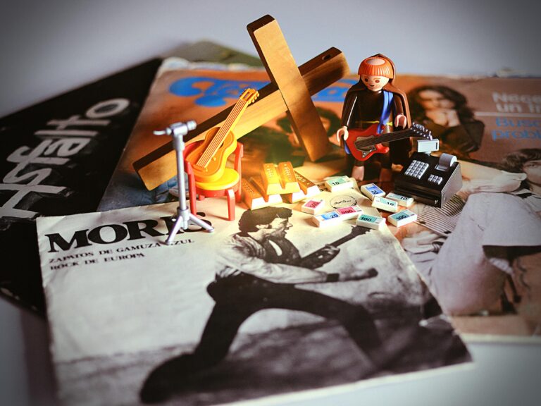 Muñeco de Playmobil con el rol de fraile con una guitarra eléctrica colgada al lado de una cruz gigante de madera. A sus pies hay billetes y lingotes de oro junto a una caja registradora. Al lado hay un micrófono en un pie y una guitarra clásica sobre una silla. Todos estos elementos están sobre discos de Moris, Tequila y Aslfalto.