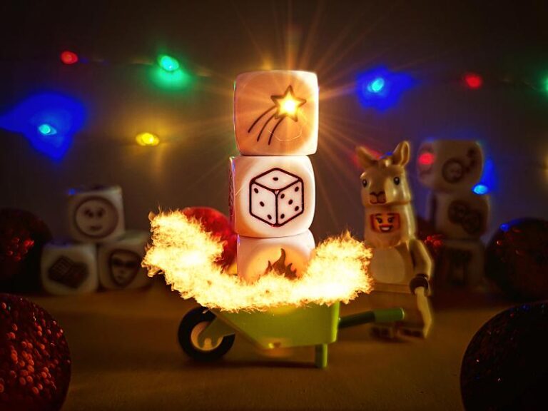 En la imagen se ven de fondo luces de navidad de colores. Una figura de lego disfrazada de llama lleva una carretilla con tres dados envueltos en llamas. En los dados se pueden ver las siguientes figuras: estrella de navidad, un dado de seis caras, una llamarada. Alrededor de la carretilla y la figura hay tiradas por el suelo bolas de navidad de color rojo y más dados con imágenes que apenas se aprecian.
