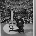 Hombre sentado en un banco en el escenario. Se ve al fondo un teatro lleno de personas.