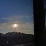 Fotografía desde una ventana. Al fondo se ven montañas y el sol en el horizonte tras una torre eléctrica.