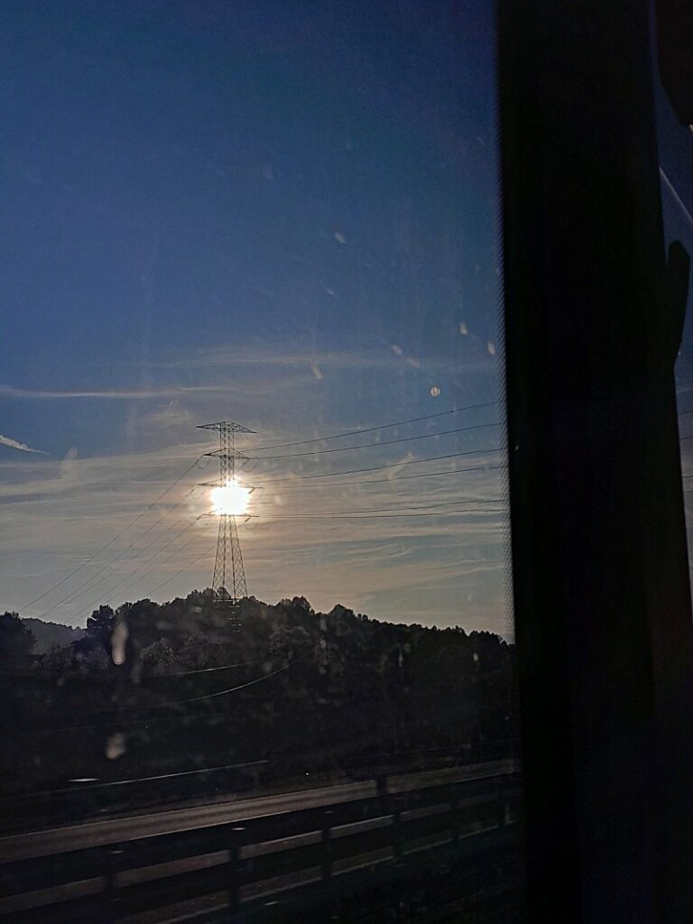 Fotografía desde una ventana. Al fondo se ven montañas y el sol en el horizonte tras una torre eléctrica.