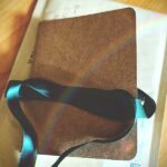 Imagen de un poemario artesanal con tapa marrón. Tiene un lazo verde alrededor ya deshecho. Está sobre una mesa de madera y un sobre de envío postal.