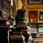 Libros amontonados sobre mesa y estanterías en lo que parece una librería o biblioteca personal.