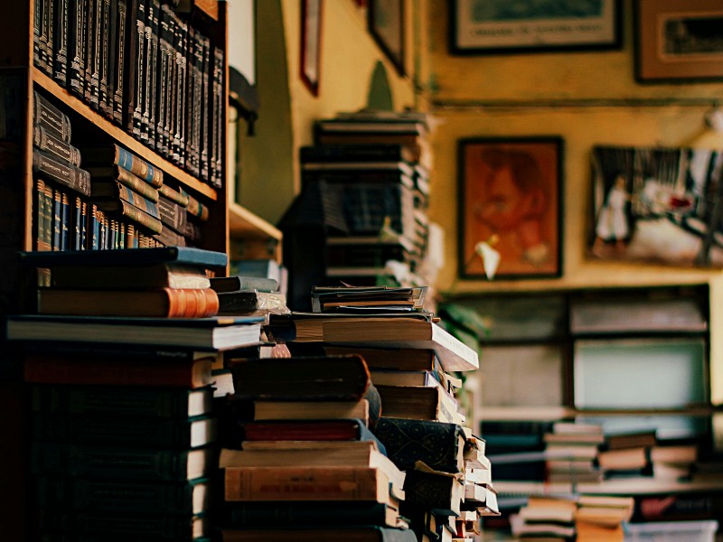 Libros amontonados sobre mesa y estanterías en lo que parece una librería o biblioteca personal.