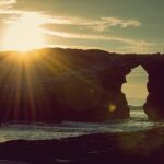 Puesta de sol en playa de As Catedrais, Ribadeo. Se ve arco de piedra por el que pasa el mar y el sol luciendo encima de la formación rocosa del arco.