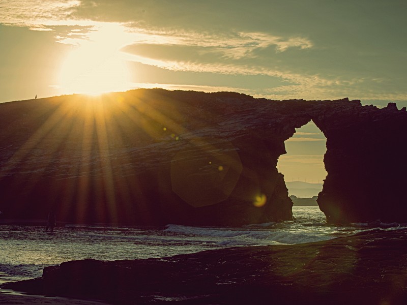 Puesta de sol en playa de As Catedrais, Ribadeo. Se ve arco de piedra por el que pasa el mar y el sol luciendo encima de la formación rocosa del arco.