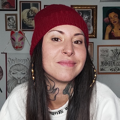 Foto color. Perfil Angi Love'n'Hate. En la imagen sale Angi con el pelo moreno, camiseta blanca y gorro rojo. Detrás de ella se ven varios cuadros.