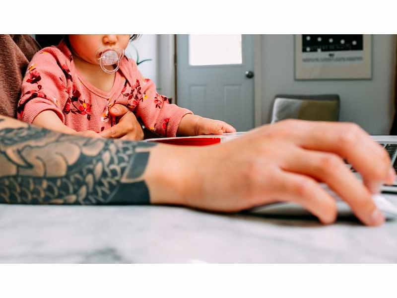 Persona con brazo tatuado que sostiene a un bebé en brazos mientras estira el brazo en acto de ir a usar el ratón de un ordenador